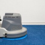 A gray floor-cleaning machine resting on bright blue carpet beside a white wall, with a blue brush underneath.