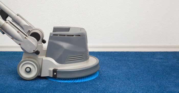 A gray floor-cleaning machine resting on bright blue carpet beside a white wall, with a blue brush underneath.