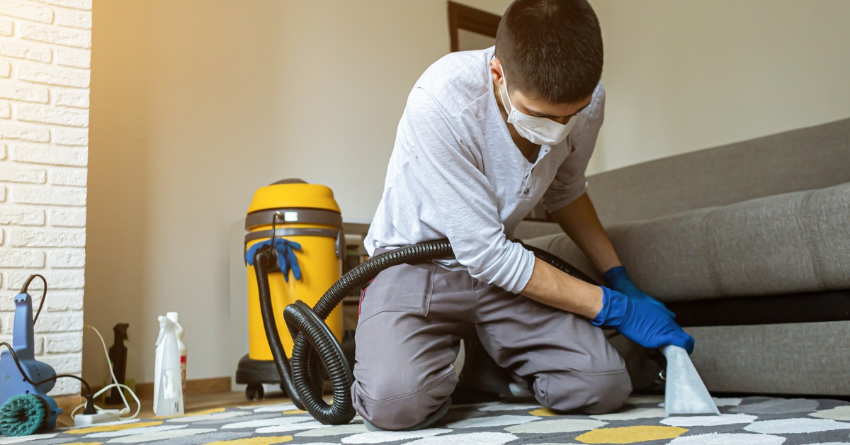 A person wearing a face mask and blue gloves kneeling on a patterned rug while using a carpet-cleaning nozzle.
