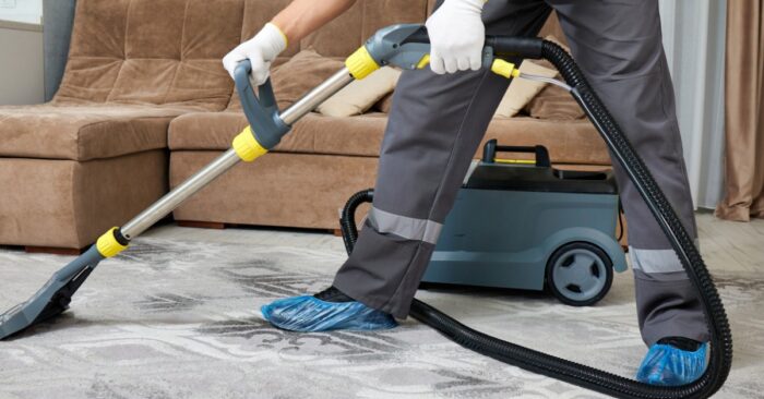 A woman in a dark uniform vacuuming a light rug beside a beige sofa in a bright living room with chairs.