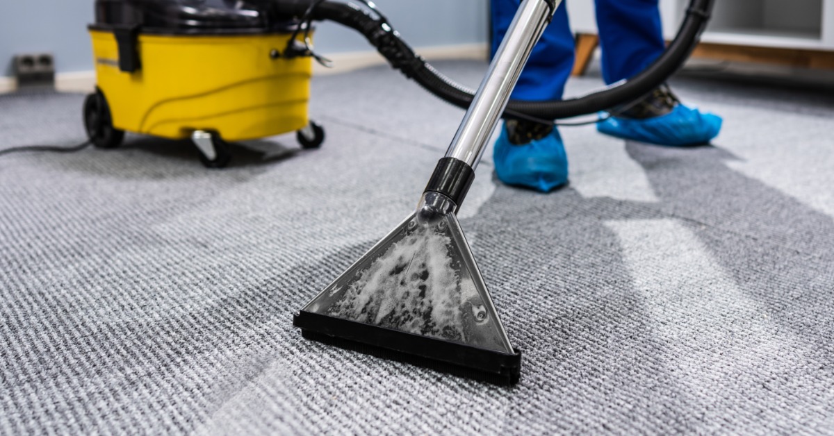 A man wearing a blue apron and yellow gloves vacuuming a light carpet near a sofa and plain white wall.
